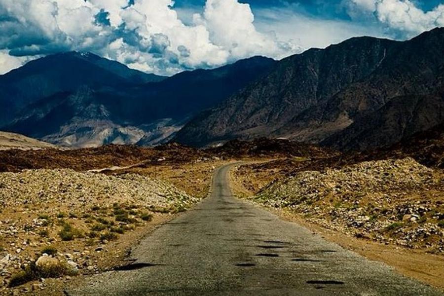 Karakoram Highway - Dirt road leading to distant mountains. A mix of short greenery and rocks on both sides of the road.