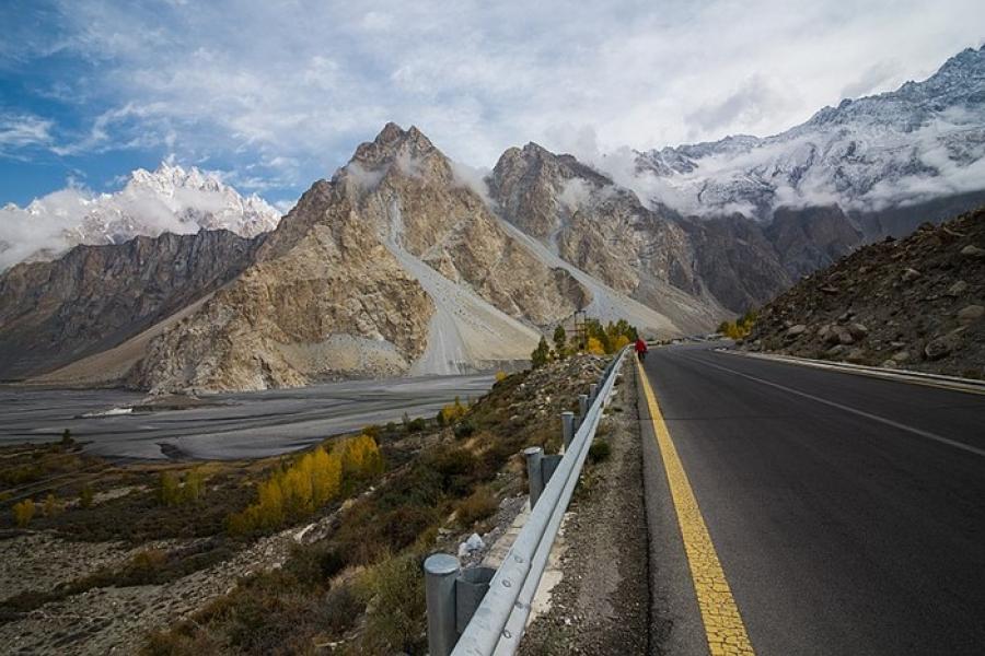 Karakoram Highway - A straight paved road in the foreground leading to craggy mountains.