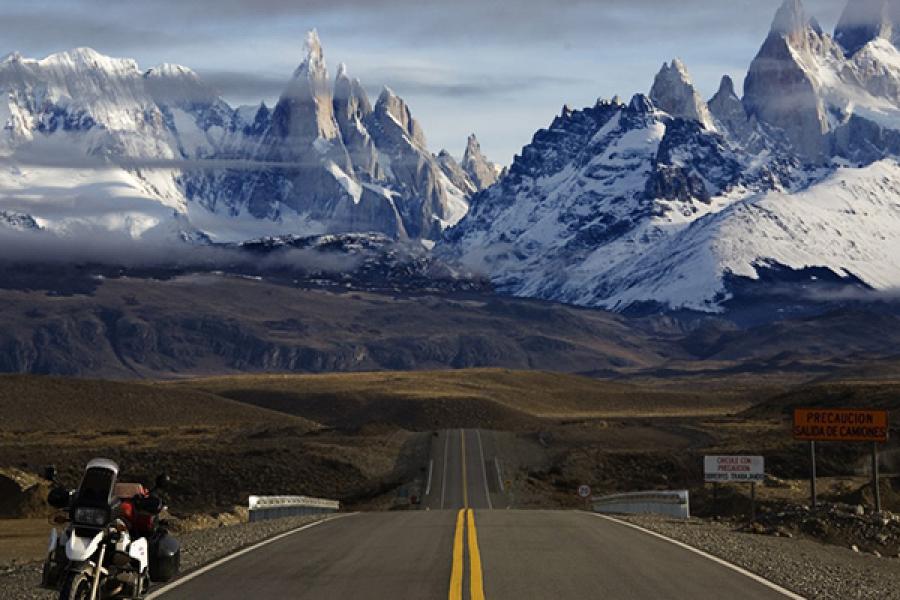 Rugged snowy mountains in the distance with a paved road in the foreground. Motorcycle parked far to the left.
