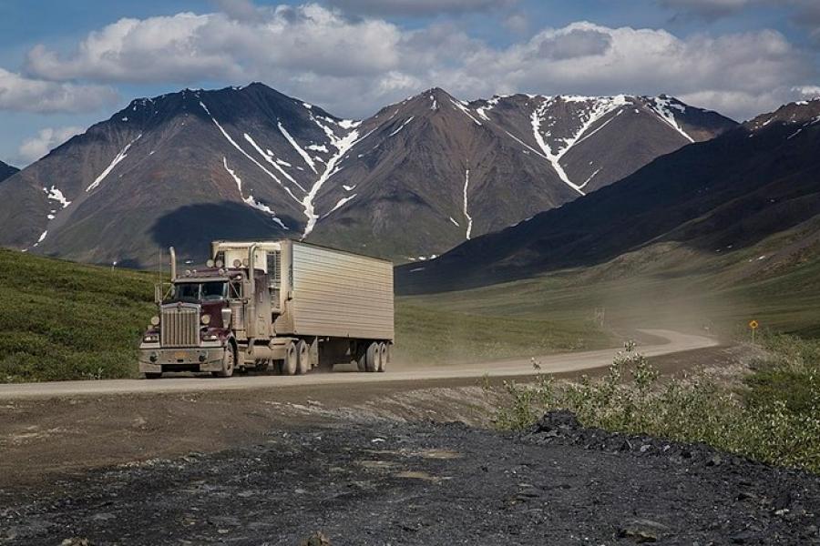 Dalton Highway - Dirt road with a transport truck kicking up dust in its wake. Mountains in the distance with traces of snow in crevices.