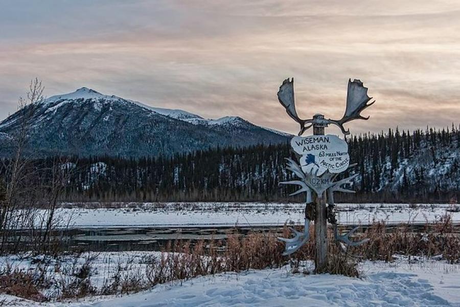 Dalton Highway - Winter dusk lighting up a grassy field partially covered in snow. A sign topped with moose antlers and decorated with other antlers. The sign says, "Wiseman, Alaska. 63 miles north of the Arctic Circle."